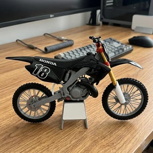 K&ouml;nnte beinhalten: Ein schwarzes und graues Honda Modellmotorrad mit der Nummer 18 auf der Seite. Das Motorrad hat goldene und rote Akzente an den Vorderradgabeln und am Lenker. Die Reifen sind schwarz mit einem detaillierten Profil. Das Modell steht auf einem kleinen wei&szlig;en St&auml;nder.