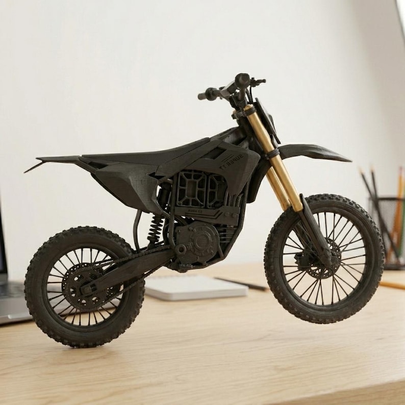 Puede incluir: Modelo de motocicleta negra con horquillas delanteras doradas. El modelo tiene un dise&ntilde;o detallado, que incluye ruedas, asiento y manillar. La motocicleta se exhibe sobre una superficie de madera, con un port&aacute;til y l&aacute;pices en el fondo.