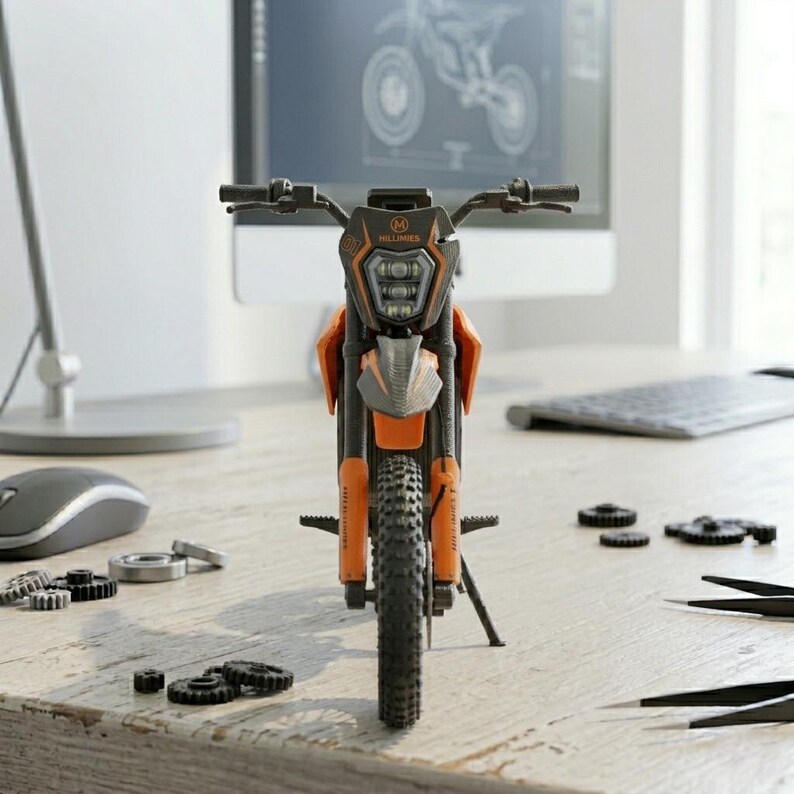 May include: An orange and black toy motorcycle with the word "HILLBILLY" on the front. The motorcycle is on a wooden desk with computer parts and a computer monitor in the background.
