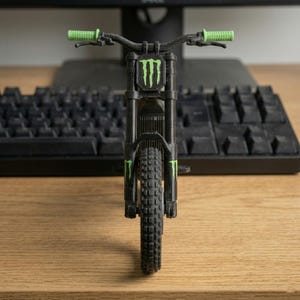 May include: A black and green miniature mountain bike model. The model features green handlebars and accents, and the Monster Energy logo on the front. The bike is positioned in front of a black keyboard on a wooden surface.