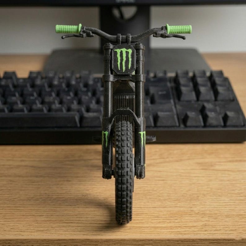 May include: A black and green miniature mountain bike model. The model features green handlebars and accents, and the Monster Energy logo on the front. The bike is positioned in front of a black keyboard on a wooden surface.