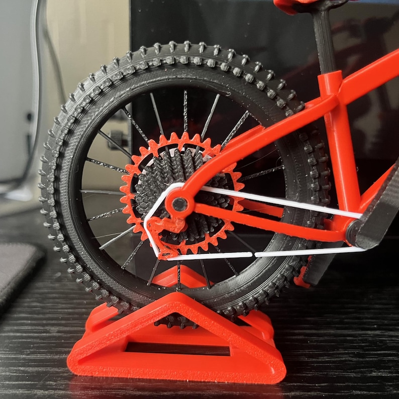 May include: A miniature red and black mountain bike model. The rear wheel has a black tire with a detailed tread pattern, silver spokes, and a red gear system. The bike is supported by a red stand.