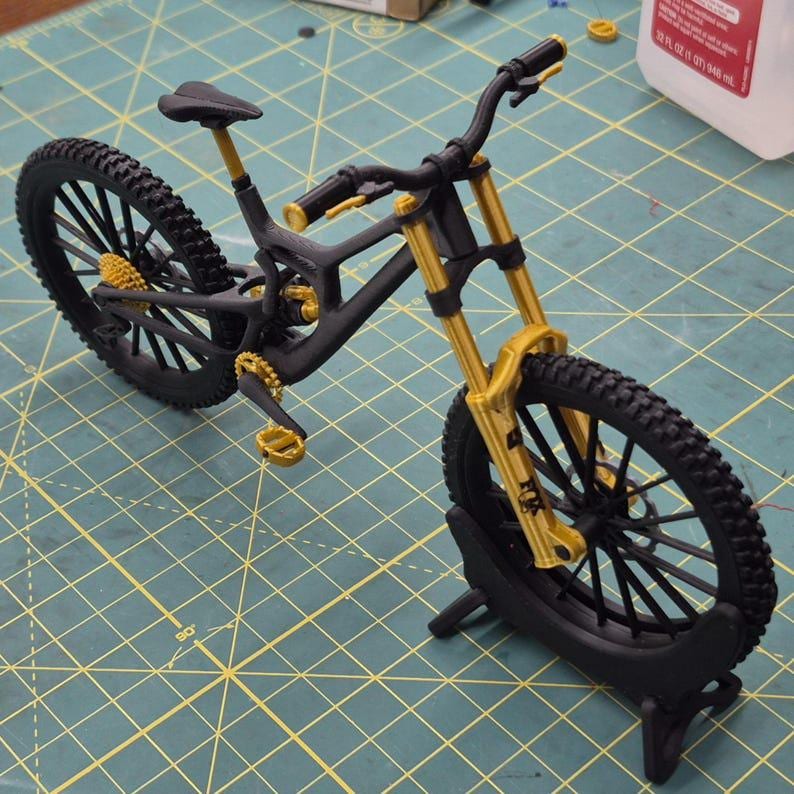 K&ouml;nnte beinhalten: Ein detailliertes Miniaturmodell eines schwarz-goldenen Mountainbikes. Das Fahrrad hat einen schwarzen Rahmen, R&auml;der und Reifen mit goldenen Akzenten an der Federung, den Pedalen und anderen Komponenten. Die Reifen haben ein Stollenprofil. Das Modell wird auf einem schwarzen St&auml;nder ausgestellt.