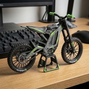 May include: A miniature black and gray motorcycle with green accents, including the handlebars and frame details. The tires have a tread pattern, and the wheels have the "Monster" logo. The motorcycle is displayed on a wooden surface.