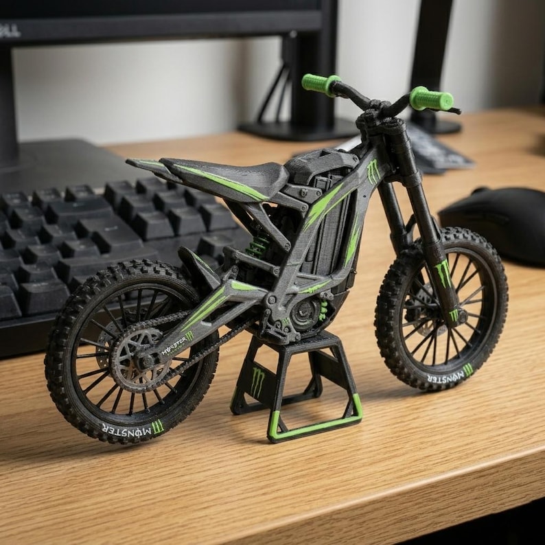 May include: A miniature black and gray motorcycle with green accents, including the handlebars and frame details. The tires have a tread pattern, and the wheels have the "Monster" logo. The motorcycle is displayed on a wooden surface.