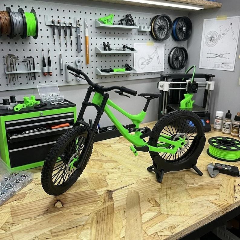 Pu&ograve; includere: Un modello di mountain bike verde lime e nero &egrave; appoggiato su un banco da lavoro in legno. La bici presenta un design a sospensione completa con pneumatici neri e un manubrio nero. Lo sfondo include strumenti, una stampante 3D e una parete forata.