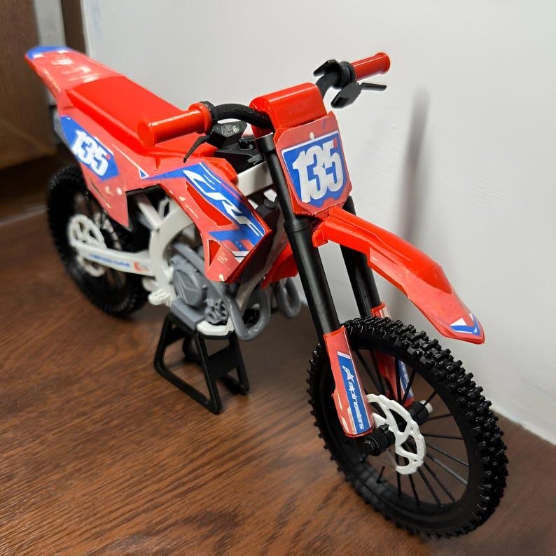 May include: A red and blue toy dirt bike with the number 135 on the front and side panels. The bike has black tires and a black stand. The handlebars and seat are red.