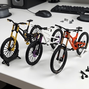 Może przedstawiać: Trzy miniaturowe modele rowerów górskich na białej powierzchni. Pierwszy jest czarny ze złotym przednim widelcem, drugi biały z fioletowymi akcentami, a trzeci pomarańczowy z napisem Santa Cruz nadrukowanym na ramie.