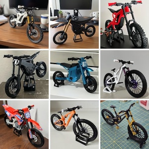 May include: A collection of miniature toy motorcycles in various colors, including white, black, red, blue, and orange. The bikes have detailed features like tires, handlebars, and seats. Some have the number 67, 87, or 135 on them.