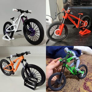 Könnte beinhalten: Eine Sammlung von Miniatur-Mountainbikes in verschiedenen Farben, darunter Weiß, Orange und Rot. Jedes Fahrrad verfügt über detaillierte Komponenten wie Federung, Reifen und Scheibenbremsen. Ein Fahrrad hat einen lila Akzent. Die Fahrräder werden auf Ständern präsentiert.
