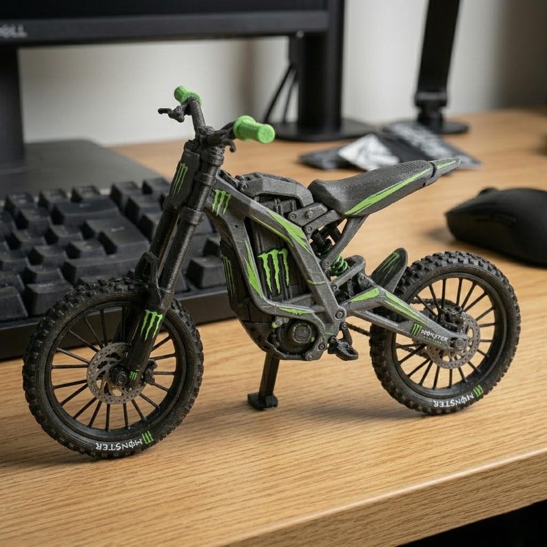 May include: A miniature black and gray motorcycle model with green accents and the "Monster" logo. The model features a detailed design, including wheels, handlebars, and a seat. The handlebars are green. The model is displayed on a wooden surface.