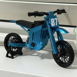 Könnte beinhalten: Ein blaues Miniaturmotorrad mit der Nummer 87 auf der Vorderseite. Das Spielzeugmotorrad hat schwarze Reifen, einen schwarzen Sitz und einen schwarzen Lenker. Das Motorrad steht auf einem schwarzen Ständer.