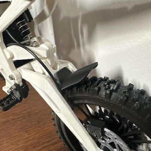 May include: Close-up of a white and black model motorcycle. The black tire has a knobby tread. The motorcycle has a black fender and a white frame. The suspension and other mechanical parts are visible.