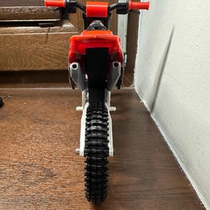 Puede incluir: Una moto de cross de juguete en miniatura roja, negra y blanca. La moto tiene un neum&aacute;tico negro con una banda de rodadura texturizada, manillar rojo y un asiento rojo. La moto est&aacute; dise&ntilde;ada para parecerse a una moto real.