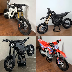 Könnte beinhalten: Vier Mini-Motocross-Motorräder in Weiß, Schwarz und Rot. Die Motorräder haben schwarze Reifen, goldene Federung und verschiedene Nummernschilder. Eines hat die Nummer 67, ein anderes 135, und die anderen haben keine sichtbaren Nummern.