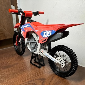 May include: A red and blue toy dirt bike with black wheels and a white frame. The number 135 is displayed on a blue background on the side of the bike. The bike is on a black stand.