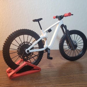 May include: A miniature white mountain bike with black tires, seat, and components. The handlebars have red grips. The bike is displayed on a red stand, showcasing its design and features. The bike is a detailed model.