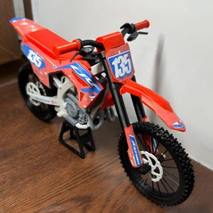 Puede incluir: Una moto de cross de juguete roja y azul, con el n&uacute;mero 135 en los paneles delantero y laterales. Tiene neum&aacute;ticos negros y un soporte negro. El manillar y el asiento son rojos.