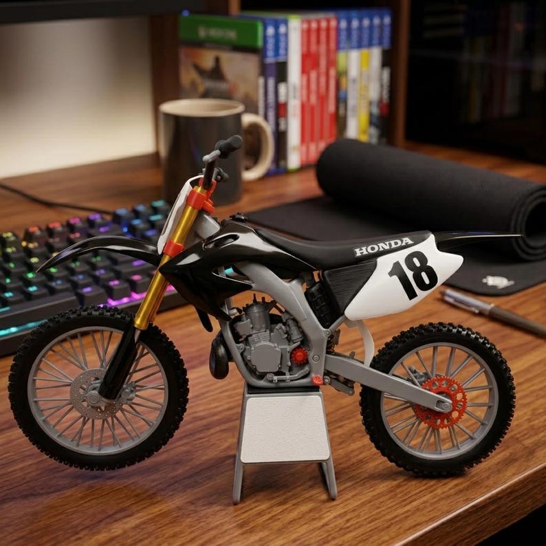 May include: A black, white, and gray Honda dirt bike model with the number 18 on the side. The model has gold front forks and red accents on the wheels. It is displayed on a wooden surface.