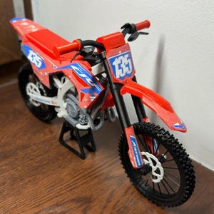 May include: A red and blue toy dirt bike with the number 135 on the front and side panels. The bike has black tires and a black stand. The handlebars and seat are red.