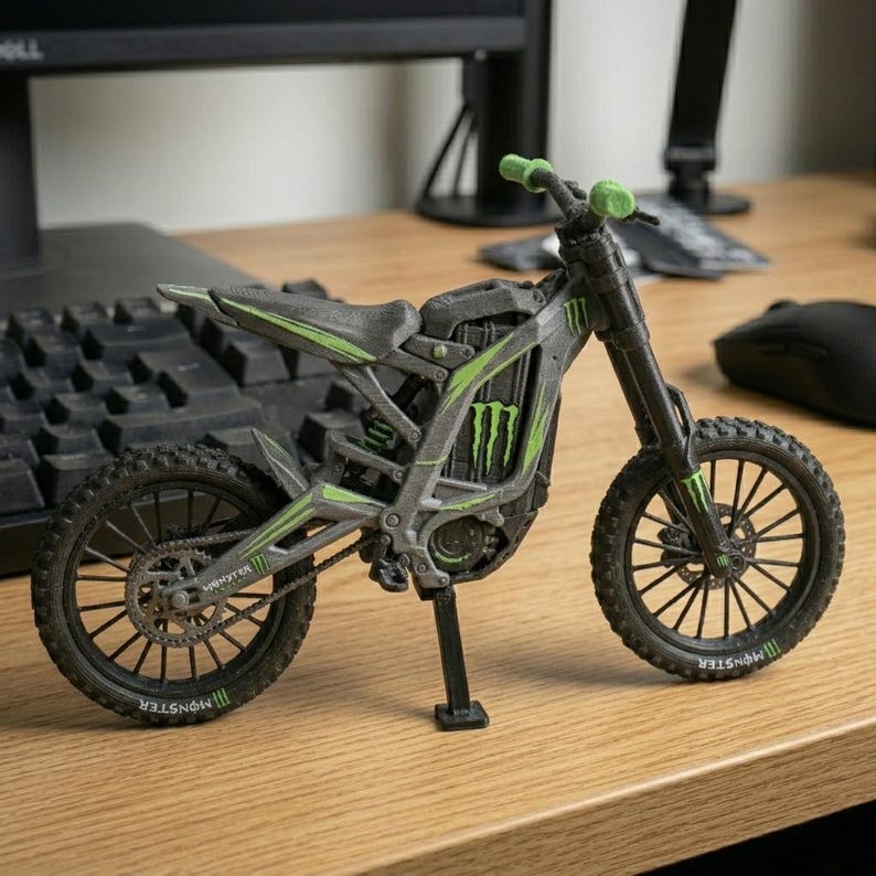 May include: A detailed model of a gray and black motorcycle with green accents. The motorcycle features the "Monster" logo and is set on a wooden surface. The handlebars and wheels are black, and the tires have a textured design.