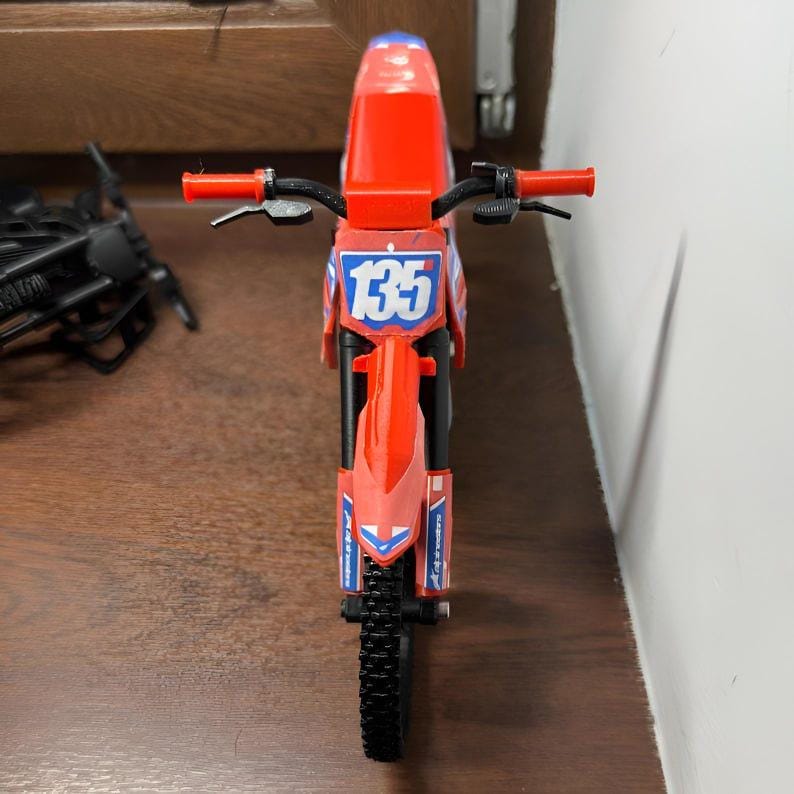 May include: A red and blue toy motorcycle with the number 135 on the front. The motorcycle has black handlebars and tires. The toy is made of plastic.