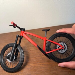 May include: A miniature red and black mountain bike model. The bike features a red frame, black tires, and a black seat. The handlebars and front suspension are also black. The bike is designed with realistic details.