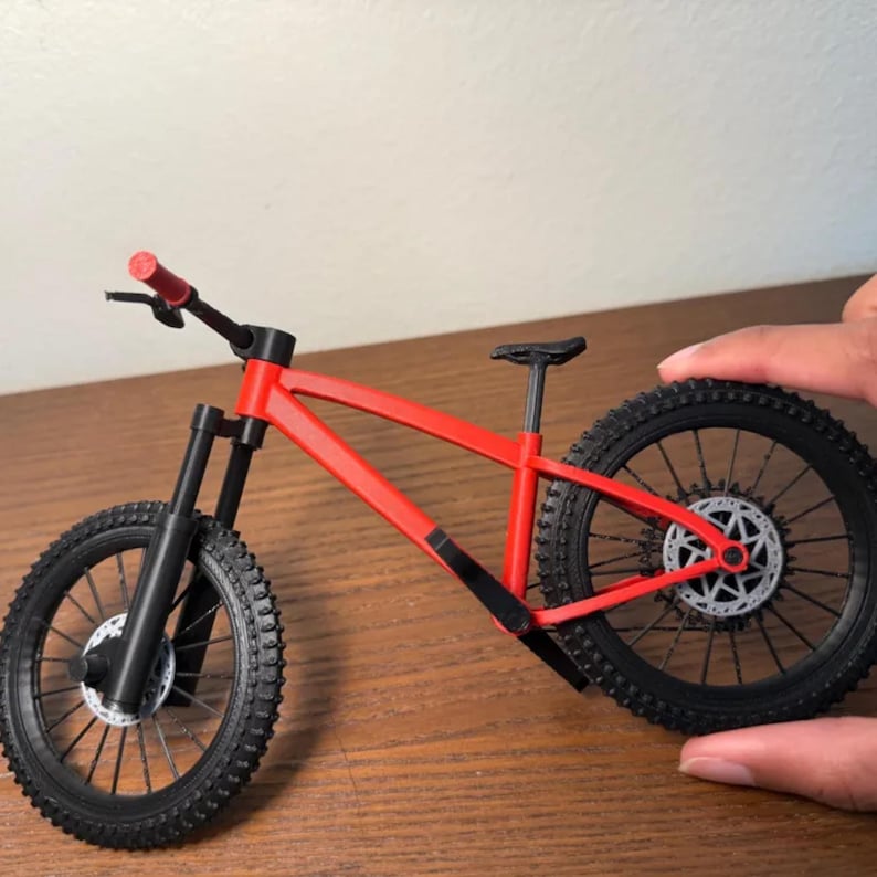 May include: A miniature red and black mountain bike model. The bike features a red frame, black tires, and a black seat. The handlebars and front suspension are also black. The bike is designed with realistic details.