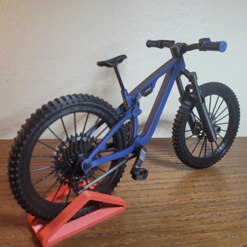 May include: A blue and black mountain bike model with a red stand. The bike has black tires, a black seat, and blue handlebars. The frame is blue, and the wheels have detailed spokes. The bike is displayed on a red stand.