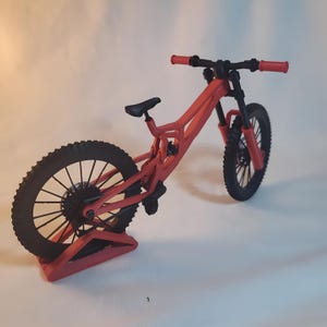K&ouml;nnte beinhalten: Ein rotes Mountainbike-Modell mit schwarzen Reifen und einem schwarzen Sattel. Der Lenker und die Vorderradaufh&auml;ngung sind rot, und das Fahrrad wird auf einem roten St&auml;nder pr&auml;sentiert. Das Fahrrad hat ein detailliertes Design.