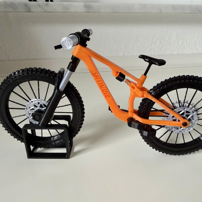 May include: An orange and black miniature mountain bike with the word "SPECIALIZED" printed on the frame. The bike has black tires, a black seat, and silver accents. It is resting on a black stand.