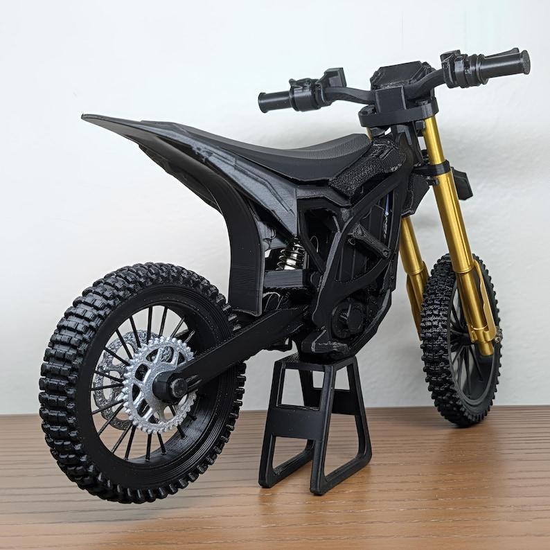 May include: A black and gold miniature motorcycle model. The motorcycle has a black frame, seat, and tires, with gold-colored front forks. The model is displayed on a black stand, showcasing the details of the design.