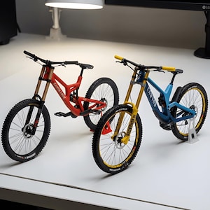 May include: Two miniature mountain bikes, one red with Santa Cruz branding and one blue with Canyon branding. Both feature black handlebars, knobby tyres, disc brakes, and suspension forks. The blue bike has yellow grips and fork legs.