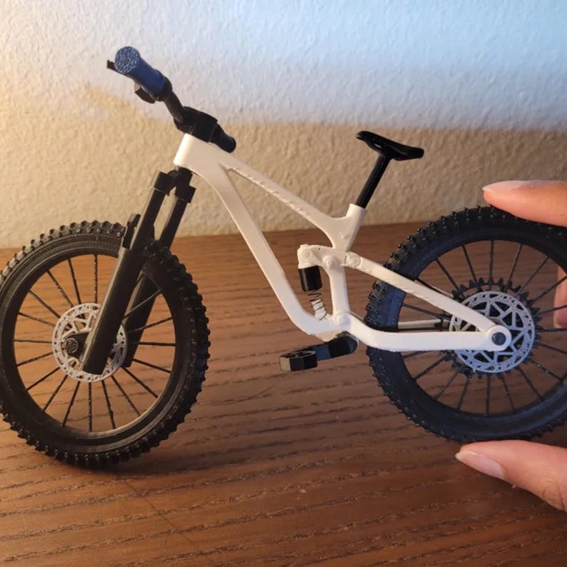 May include: A miniature white and black mountain bike with blue handlebar grips. The bike has black tires, a black seat, and a detailed frame. The bike is held by a hand.
