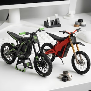 Op de afbeelding: Twee miniatuur elektrische crossmotor-modellen. De ene is zwart met groene accenten en Monster Energy-branding, gemonteerd op een zwart-groene standaard. De andere is rood met zwarte accenten en goudkleurige voorvorken. Beide hebben noppenbanden.
