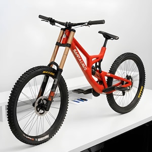 Könnte beinhalten: Ein leuchtend oranges Santa Cruz Mountainbike mit vollgefedertem Rahmen, schwarzem Lenker, schwarzem Sattel und schwarzen Stollenreifen mit Maxxis-Schriftzug. Die Vorderradgabel hat goldfarbene Standrohre und ein schwarzes Federungssystem.