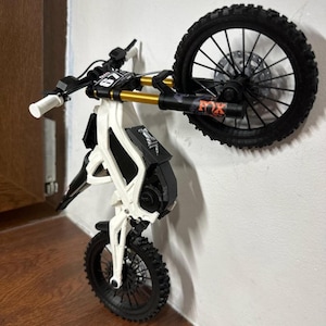 May include: A white and black miniature dirt bike model with a black tire and gold suspension. The model is mounted on a white wall. The front tire is on the ground, and the rear tire is raised. The model has the text "FOX" on the suspension.