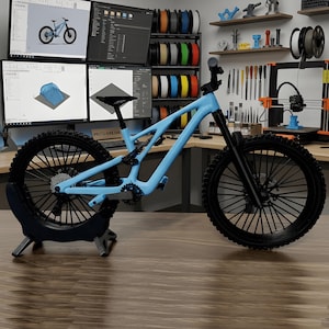 Puede incluir: Bicicleta de monta&ntilde;a azul claro con neum&aacute;ticos y componentes negros. La bicicleta se exhibe sobre una superficie de madera, con monitores de computadora y equipos de impresi&oacute;n 3D en el fondo. Es un modelo de suspensi&oacute;n completa.
