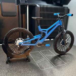 Pu&ograve; includere: Una mountain bike blu in miniatura con pneumatici neri e sella nera. La bici ha manubrio e pedali blu. La ruota posteriore &egrave; montata su un supporto nero. La bici &egrave; un modello dettagliato.