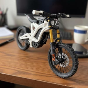 May include: A detailed model of a white and black electric dirt bike with gold suspension forks. The bike features black tyres, a number plate with the number 67, and the word "GUTS". The model is displayed on a wooden surface.