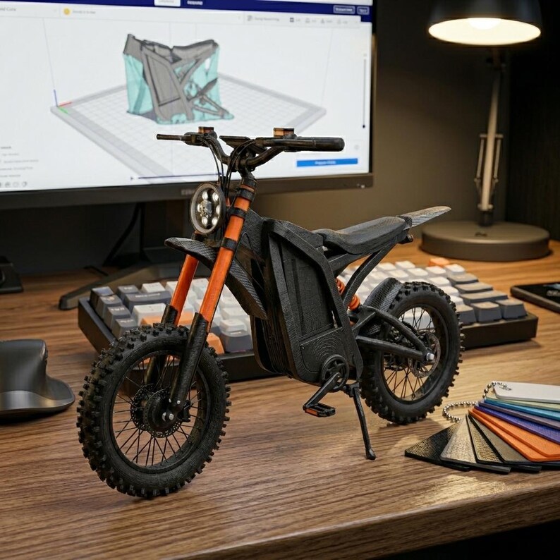 K&ouml;nnte beinhalten: Ein schwarzes und orangefarbenes Miniaturmotorradmodell mit Stollenreifen, Scheinwerfer und Sitz. Das Modell steht auf einer Holzoberfl&auml;che, im Hintergrund ein Computermonitor mit einem 3D-Design.