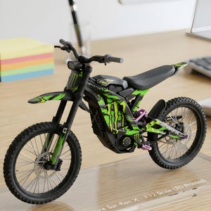 May include: A 1:12 scale 3D printed model of a Sur-Ron X electric dirt bike. The model features a black frame with bright green graphics, a black seat, knobby tyres, and the Monster Energy logo on the side panels and front mudguard.