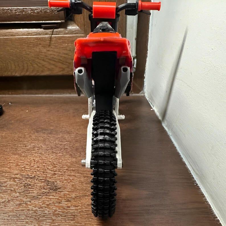 Puede incluir: Una motocicleta de juguete con un marco rojo y negro, neum&aacute;ticos negros y manillar rojo. La motocicleta tiene un dise&ntilde;o detallado, incluyendo tubos de escape y una banda de rodadura texturizada. La imagen est&aacute; tomada desde un &aacute;ngulo bajo, mostrando la parte trasera del veh&iacute;culo.