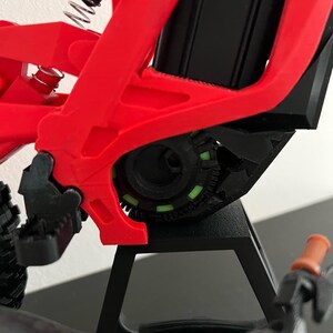 May include: Close-up of a red and black electric bicycle frame. The frame has a complex design with a black motor housing and a green and black gear system. The handlebars are orange.