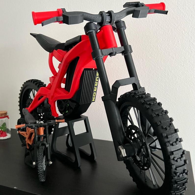 K&ouml;nnte beinhalten: Ein rotes und schwarzes Surron Elektro-Dirtbike-Modell mit schwarzen Reifen und Lenker. Das Modell wird auf einem schwarzen St&auml;nder ausgestellt. Ein kleineres orangefarbenes und schwarzes Modell befindet sich vor dem gr&ouml;&szlig;eren.