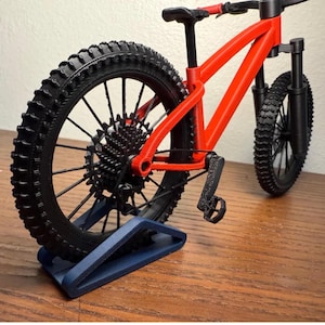 May include: A miniature red mountain bike model with black tires and components. The bike is displayed on a blue stand, showcasing the intricate details of the frame, wheels, and gears. The tires have a textured tread.