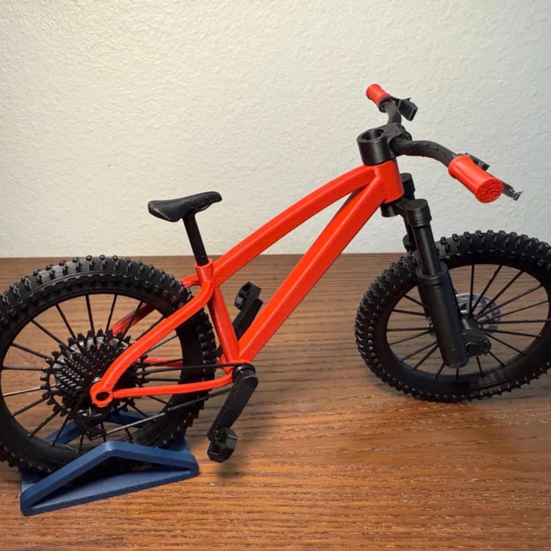 May include: A miniature red mountain bike with black tires and components. The bike has a black seat and handlebars with red grips. The bike is displayed on a blue stand, showcasing its design and features. The tires have a knobby tread.