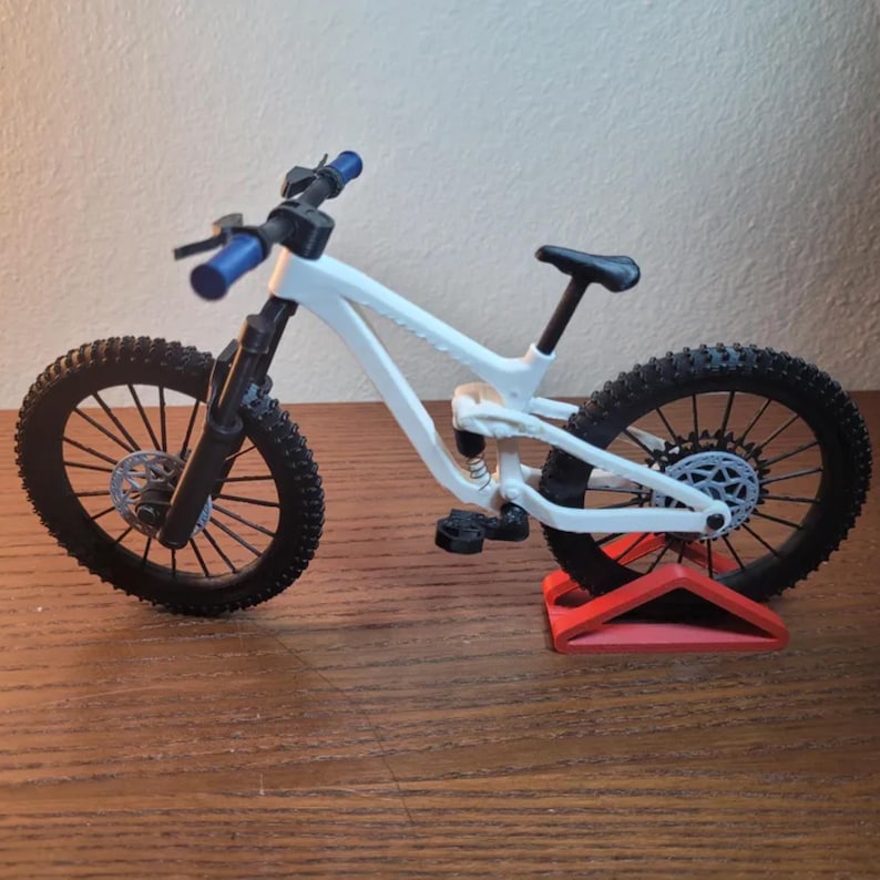 May include: A white and black mountain bike model with blue handlebars and black tires. The bike is displayed on a red stand, showcasing its intricate design and details. The wheels have a detailed spoke pattern.