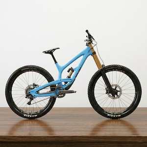 May include: A light blue mountain bike with black tires and a black seat. The bike has gold-colored suspension forks and a black handlebar. The tires have the word "MAXXIS" printed on them. The bike is on a wooden surface.