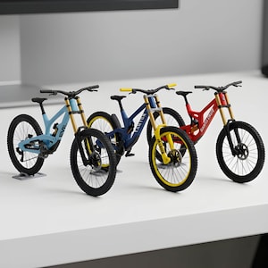 Puede incluir: Tres modelos de bicicleta de montaña en miniatura dispuestos en fila. La primera es azul claro con detalles negros, la segunda es azul oscuro con manillar y llanta delantera amarillos, y la tercera es roja con componentes negros. Tienen horquillas doradas.